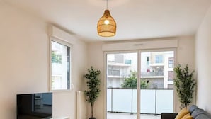 Un salon moderne avec un canapé, une table basse, un téléviseur posé sur un meuble et un balcon agrémenté de plantes.