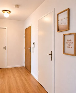 A hallway with wooden flooring, white walls, and two doors, one with a wooden panel and the other with a white panel. There are two framed pictures on the wall. - Brand New Family Apartment, Terrace & Parking, 15 min from Disneyland (Bussy-Saint-Georges)