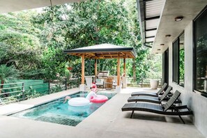 Ein Pool mit Liegestühlen, einer Laube und Blick auf üppiges Grün.