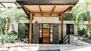 Un bâtiment moderne doté d’une pergola en bois à l’entrée, d’un mur en pierre et d’un chemin en béton menant à la porte.