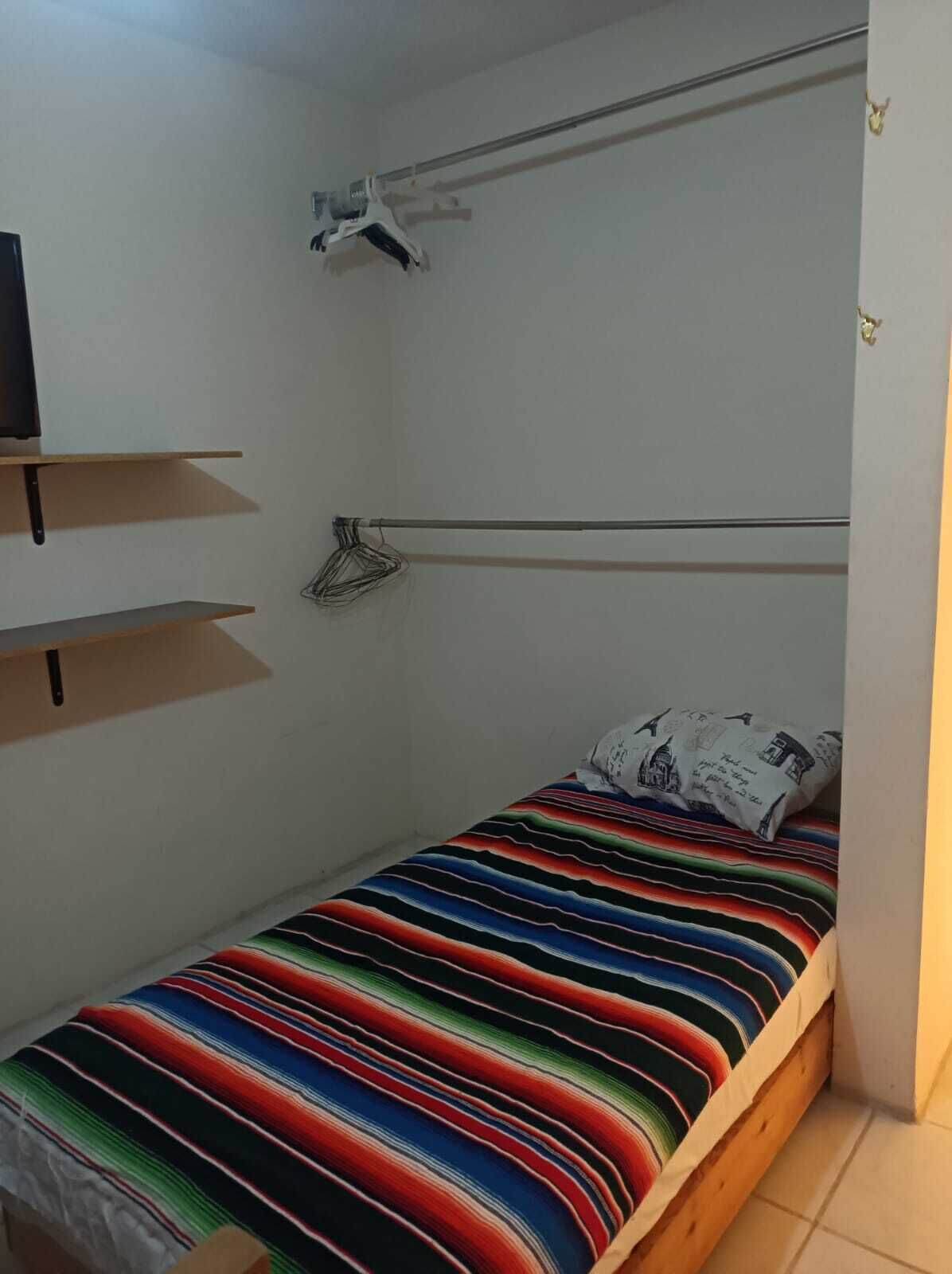 Een kleine eenpersoonskamer met een kleurrijk gestreept dekbed, een plafondventilator en een wandplank.