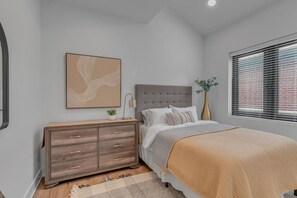 Ein Schlafzimmer mit einem Bett, einem Schminktisch, einem Nachttisch, einer Lampe, einer Pflanzvase, einem Gemälde und einem Fenster mit Jalousien.
