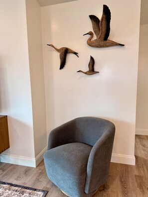 Una habitación con un sillón gris, piso de madera y esculturas de pájaros en la pared.