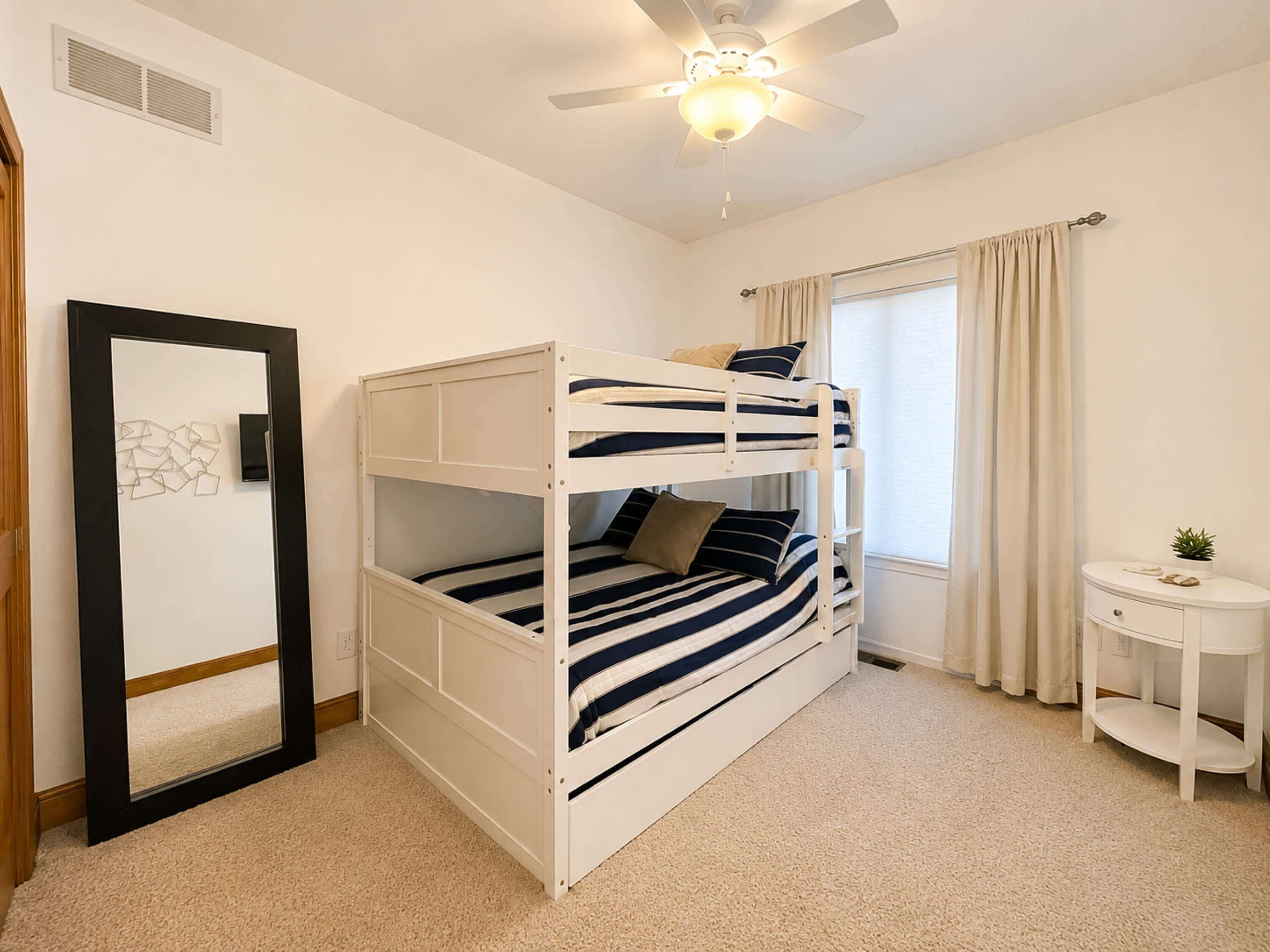Une chambre avec un lit superposé, un ventilateur de plafond, une fenêtre avec des rideaux et une petite table de chevet agrémentée d’une plante.