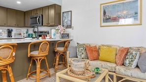 Eine Küche mit Holzausstattung, einer Mikrowelle und einer Kaffeemaschine. Es gibt einen Essbereich mit Korbstühlen und ein Sofa mit gemusterten Kissen.