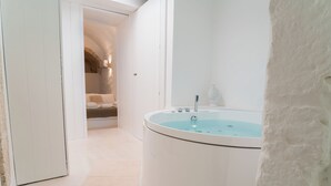 Un baño moderno con una bañera independiente, una puerta blanca y una habitación visible con una cama y una mesita de noche.