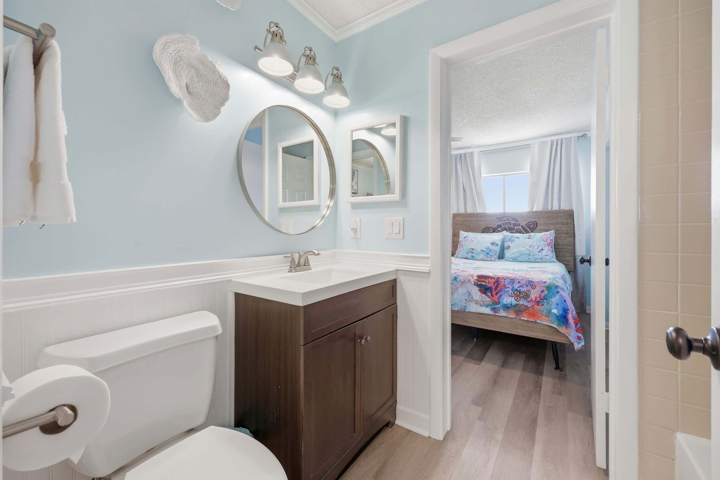 A bathroom with a toilet, sink, and mirrors. There is a bed with a colorful bedspread in the adjacent room.