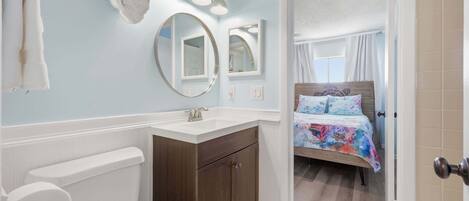 A bathroom with a toilet, sink, and mirrors. There is a bed with a colorful bedspread in the adjacent room.