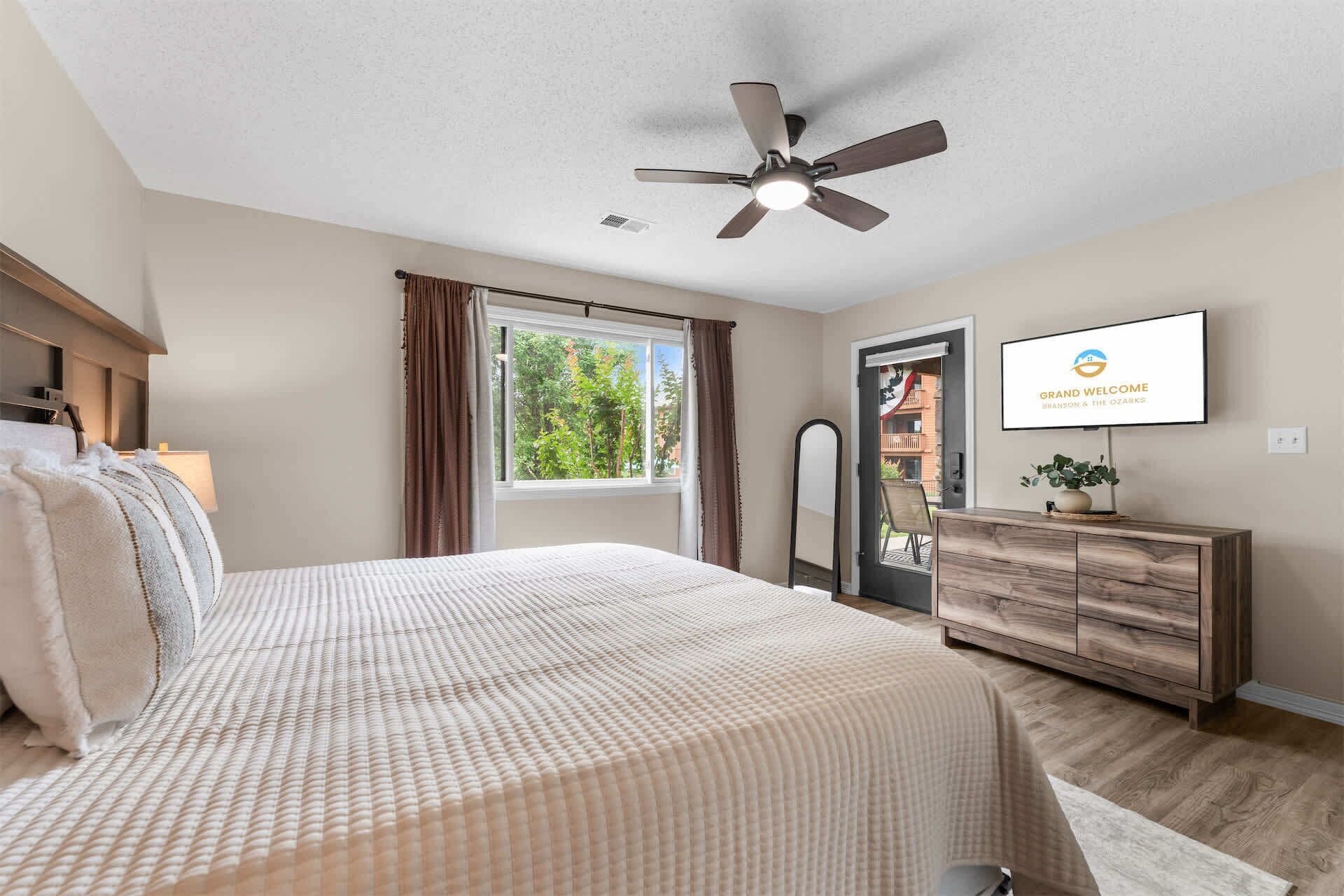 A bedroom with a bed, a ceiling fan, a window with curtains, a dresser, and a TV displaying 'Grand Welcome'.