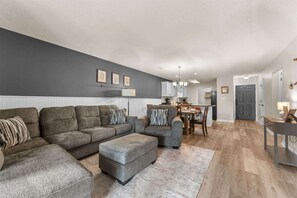 Ein modernes Wohnzimmer mit einer grauen Sofagarnitur, einem Essbereich und einer Küche im Hintergrund.