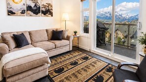 Een woonkamer met een bank, een vloerkleed, een lamp en een uitzicht op bergen door grote ramen.