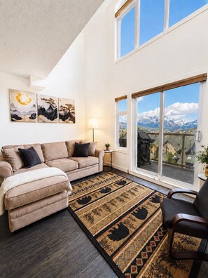 Ein Wohnzimmer mit einer Couch, einem Teppich, einer Lampe und Blick auf Berge durch große Fenster.