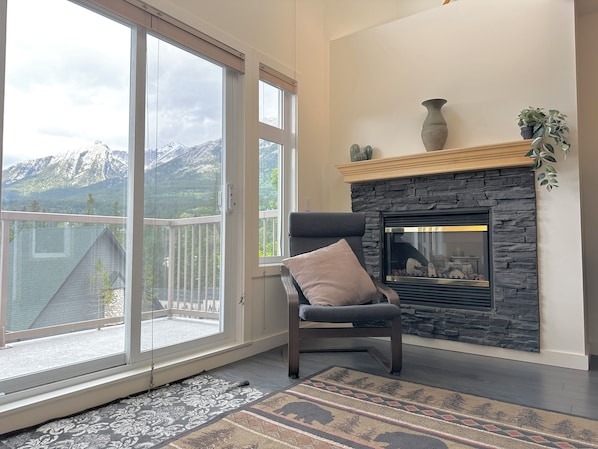 Ein Zimmer mit Kamin, einem Sessel, einem Teppich und Blick auf Berge durch große Fenster.
