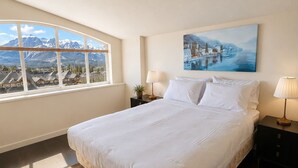 Een slaapkamer met een groot bed, een raam met uitzicht op bergen, een schilderij aan de muur en een nachtkastje met een lamp.
