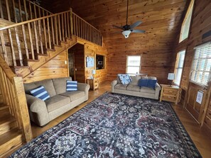A log cabin interior with a staircase, two sofas, a patterned rug, and a ceiling fan.