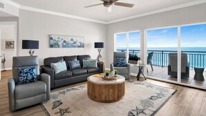 A modern living room with a large window, a ceiling fan, and a view of the ocean. - Beachfront 2BR @ Admiral Quarters • Sleeps 8 • Stunning Views (Orange Beach)