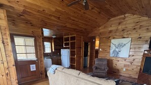 Interno di una baita in legno con un divano, una poltrona, una ventola a soffitto e un frigorifero.