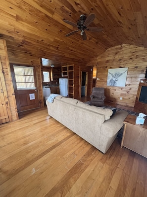 A wooden cabin interior with a sofa, a chair, a ceiling fan, and a refrigerator.
