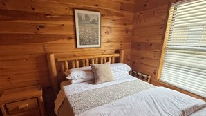 Camera da letto in una baita di legno, con un letto, un comodino, una cassettiera, una ventola a soffitto e una finestra con persiane.