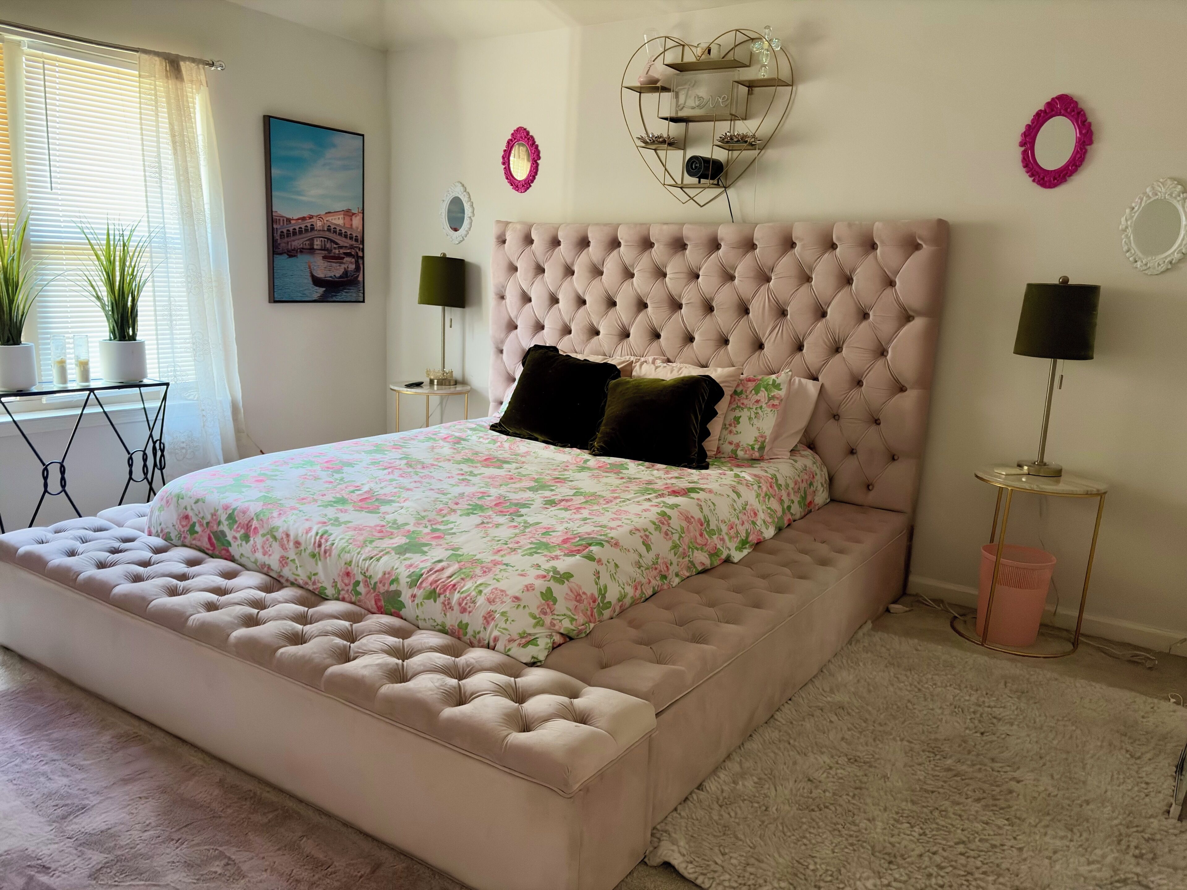 A bedroom with a tufted headboard, floral bedding, a heart-shaped wall decoration, and a framed picture of a canal.