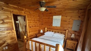 Ett sovrum i en stuga med träpanel, inrett med en säng, sängbord, en takfläkt och en dörr som leder till ett annat rum.