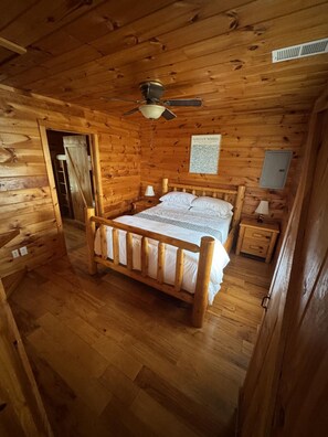 A wooden cabin bedroom with a bed, nightstands, a ceiling fan, and a door leading to another room.