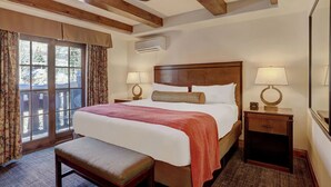 Habitación de hotel con una cama grande, un banco, una mesita de noche con lámpara, una ventana con cortinas y un equipo de aire acondicionado.