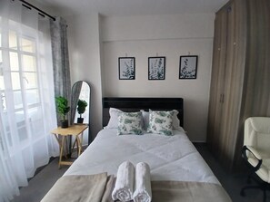 A hotel room with a bed, a desk, a chair, a mirror, and three framed pictures on the wall. - Elegant Studio Apartment for Business & Leisure (Nairobi)