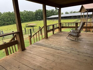 A wooden deck with a rocking chair, a fenced pasture, and a barn in the background.