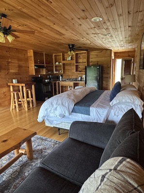 A log cabin interior with a bed, sofa, wooden dining table, kitchen area, and a ceiling fan.