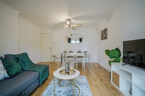 A living room with a sofa, a dining table with chairs, a ceiling fan, a mirror, and a TV on a stand.