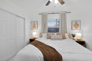 Un dormitorio con cama, dos mesitas de noche con lámparas, ventana con cortinas, ventilador de techo y puerta.