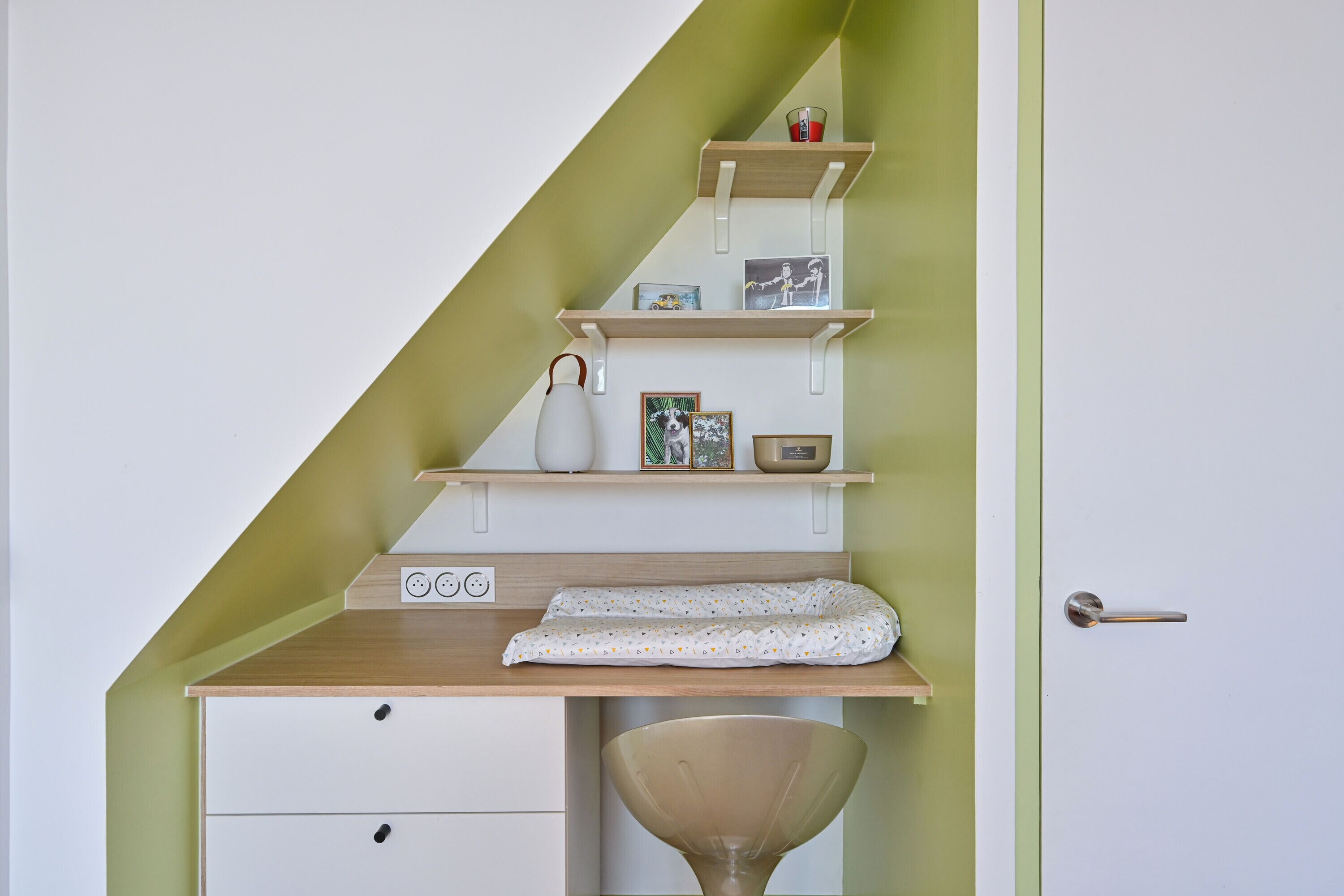Una stanza con una scrivania in legno, una sedia bianca, un piccolo ripiano con oggetti decorativi e una porta con maniglia.