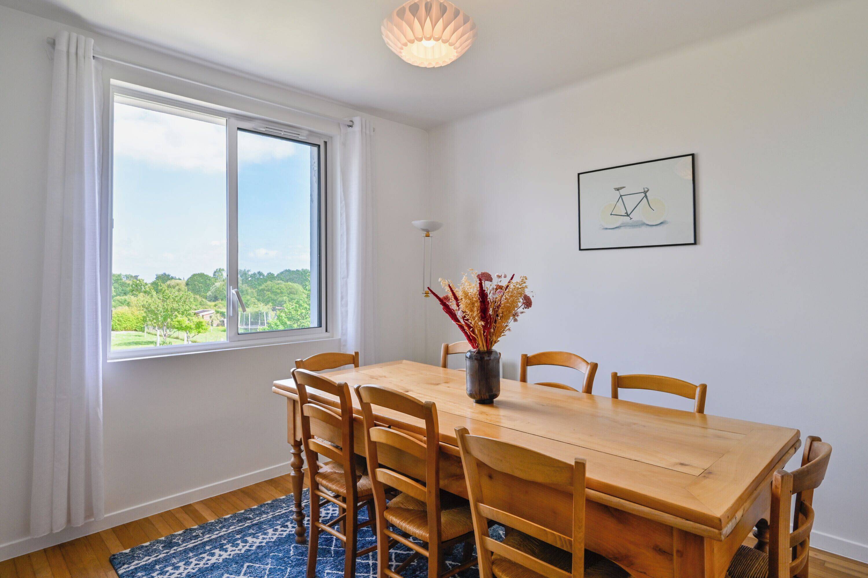 A dining area with a wooden table and chairs, a vase with dried flowers, a framed picture on the wall, and a view through a large window.