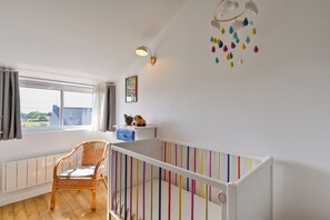 Et værelse med en rejsebarneseng, en fletstol, et vindue med gardiner, en vægmonteret lampe og en farverig uro.