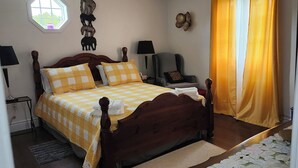 Una cama bien hecha con una colcha a cuadros, una mesita de noche con lámpara, una silla y una ventana con cortinas.