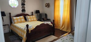 Ein ordentlich bezogenes Bett mit kariertem Überwurf, ein Nachttisch mit Lampe, ein Stuhl und ein Fenster mit Vorhängen.
