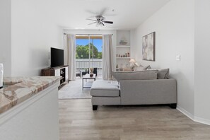 A modern living room with a grey sofa, a flat-screen TV, a ceiling fan, and a view of a balcony with a railing. - Comfy & Modern Apt w/ Themed Room | Near Disney (8908-203) (Kissimmee)
