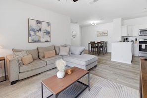 Ein modernes Wohnzimmer mit einem grauen Sofa, einem Holztisch und einer Küche im Hintergrund.