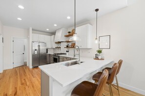 A modern kitchen with white cabinetry, stainless steel appliances, a central island with bar stools, and pendant lighting. - Old Town North Retreat Bikes, Brews & Trails (Fort Collins)