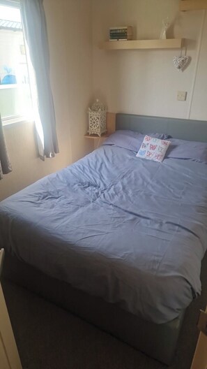 A bedroom with a bed, a nightstand, a wall-mounted shelf, and a window with curtains. - Coastfields 8 Berth Holiday Home (Skegness)