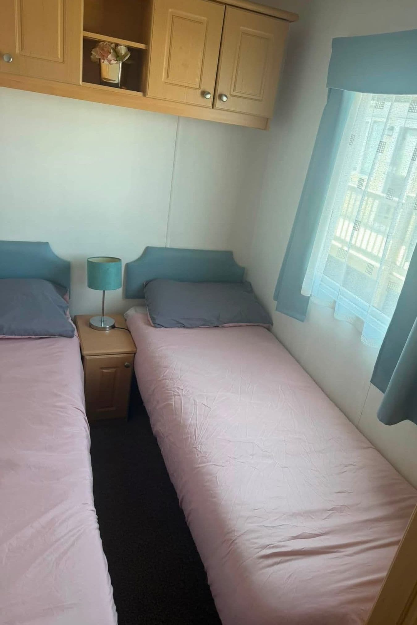 Un dormitorio con dos camas individuales, una mesita de noche con lámpara y una ventana con vistas a edificios.