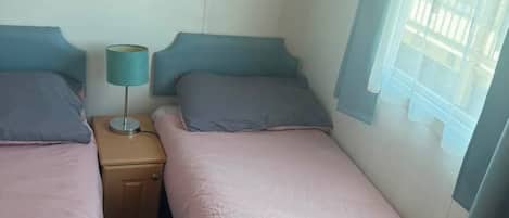 Un dormitorio con dos camas individuales, una mesita de noche con lámpara y una ventana con vistas a edificios.
