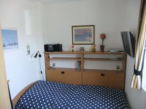Ein Zimmer mit einem Bett, einem Schreibtisch mit Computer, einem gerahmten Bild an der Wand und einem Fenster.
