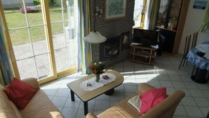 En stue med sofa, lænestol, fjernsyn, pejs og udsigt til haven gennem store vinduer.