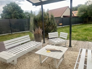 En udendørs terrasse med to hvide bænke, et bord og en potteplante.