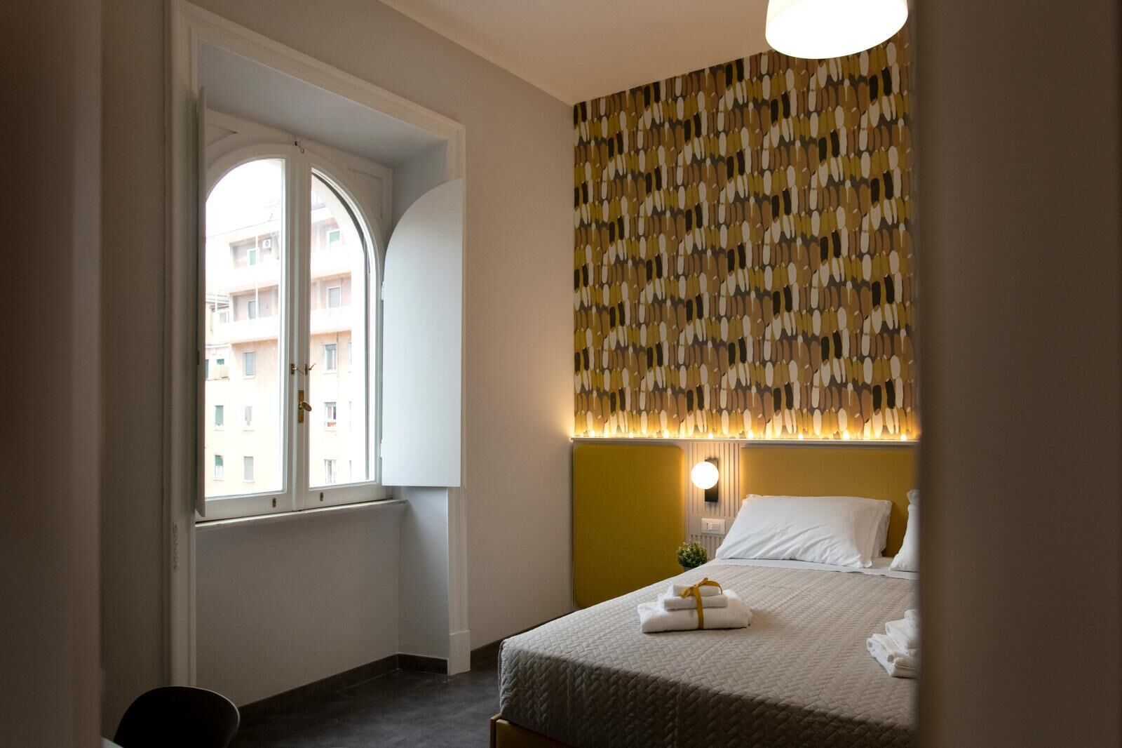 A hotel room with a bed, a window, a patterned wall, and a wall-mounted light.