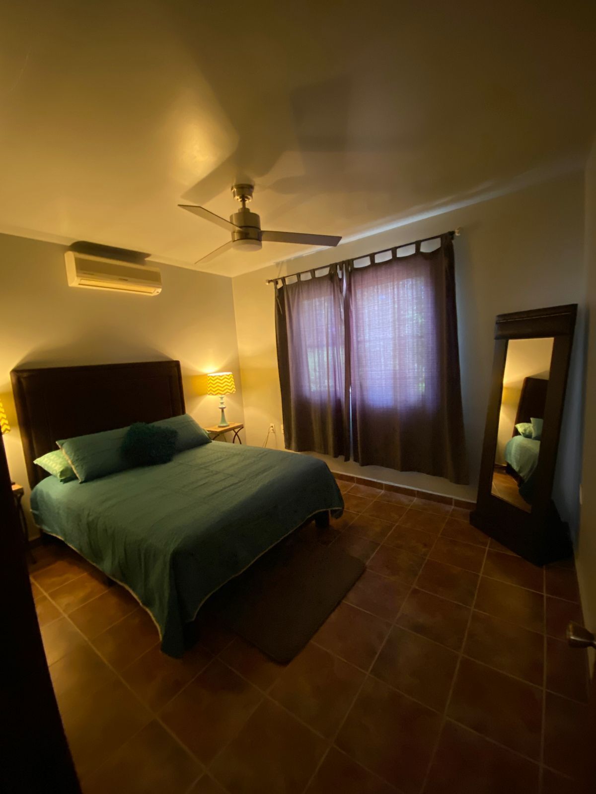 Een slaapkamer met een bed, een plafondventilator, een raam met gordijnen en een spiegel.