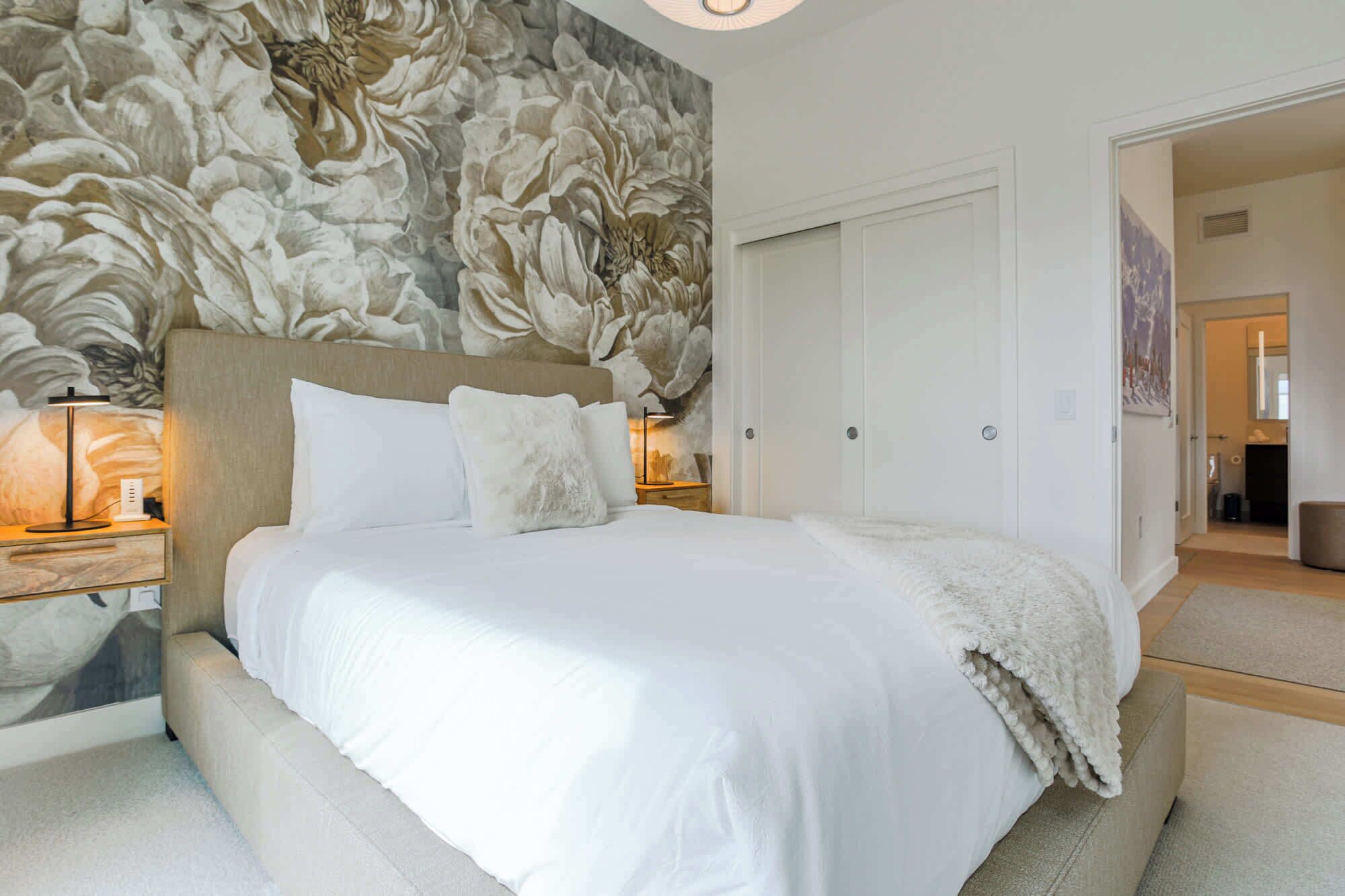 A bedroom with a large bed, white bedding, a headboard with a floral mural, two bedside lamps, and a white wardrobe.