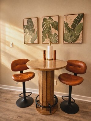 Une table ronde en bois, sur laquelle est posé un vase blanc, est entourée de deux fauteuils en cuir marron dont les pieds sont en métal noir. Le tout est placé devant un mur beige orné de trois tableaux encadrés représentant des feuilles tropicales.